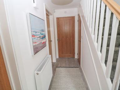 A hallway with wooden doors a radiator a painting on the wall a rug on the floor and stairs with white banisters at Summer Rose in Craster