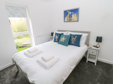 A bedroom with a bed with white linens and decorative pillows a nightstand with a lamp and a window at Summer Rose in Craster