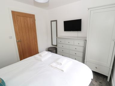 A bedroom with a bed, wooden door, wall-mounted TV, dresser, wardrobe, mirror, and folded towels on the bed at Summer Rose in Craster