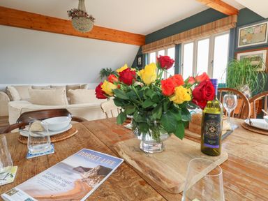 A dining table with a vase of red yellow and orange roses a wine bottle and a magazine in a room with beige sofas and large windows at Sandy Combe in West Bay