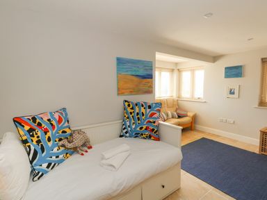 A room with a daybed with colorful pillows and folded towels a small sofa with cushions paintings and a blue rug at Sandy Combe in West Bay
