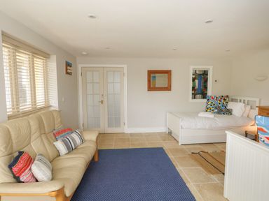 A room with a beige leather sofa with cushions a single white bed with colorful pillows a blue rug tiled floor and double glass doors at Sandy Combe in West Bay
