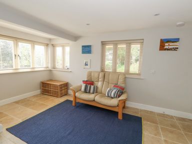 A sitting room with a beige loveseat on a blue rug and windows with blinds at Sandy Combe in West Bay