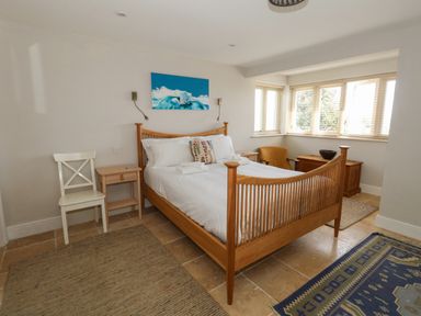 A bedroom with a wooden bed white bedding two bedside tables a white chair a mustard armchair and windows with blinds at Sandy Combe in West Bay