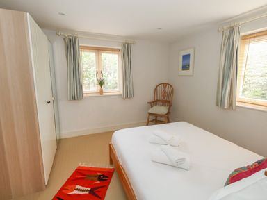 A bedroom with a bed, two windows with curtains, a wooden chair, a wardrobe, and a red rug at Sandy Combe in West Bay