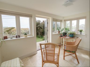 A sunroom with rattan chairs a table with plants and an open door leading outside at Sandy Combe in West Bay