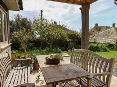 A wooden outdoor table with two wooden benches under a covered patio overlooking a garden with trees and plants at Sandy Combe in West Bay