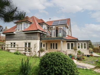 A two-story house with red tiled roof solar panels and a balcony surrounded by a garden at Sandy Combe in West Bay