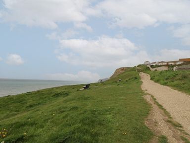 A grassy coastal path with benches overlooking the sea and houses on a hill at Sandy Combe in West Bay