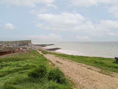 A coastal pathway with grass on both sides leading to a beach and sea with buildings and cliffs in the background at Sandy Combe in West Bay