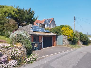 A house with solar panels on the roof and a garage next to a road with flowers and greenery at Sandy Combe in West Bay