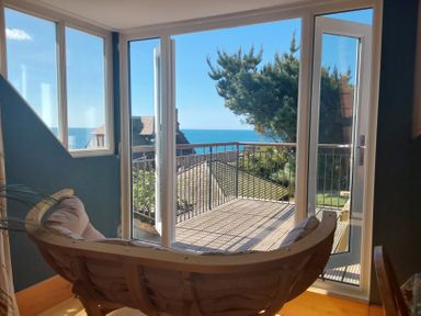 A room with a curved wooden chair facing glass doors leading to a balcony with a sea view at Sandy Combe in West Bay