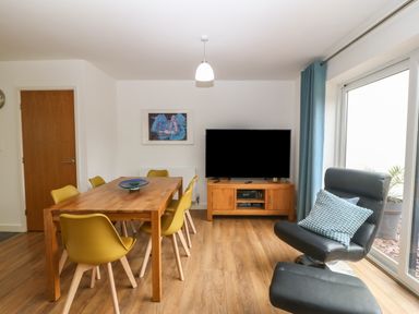A dining area with a wooden table and yellow chairs next to a TV on a wooden stand and a black armchair with a blue cushion at Hen Gapel in Waunfawr Gwynedd