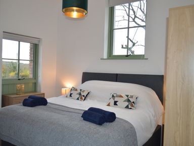 A bedroom with a double bed, two windows, bedside lamp, wooden dresser, and rolled towels at Cwrt y Wennol in Salem near Clydach