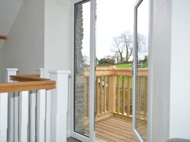 An open glass door leading to a wooden balcony with railing and a view of trees and grass at Cwrt y Wennol in Salem near Clydach
