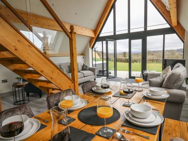 A dining table set with glasses of orange juice and wine next to a living area with sofas and large windows overlooking a garden at Hill Radnor - The Sheepfold in Llanyre near Llandrindod Wells