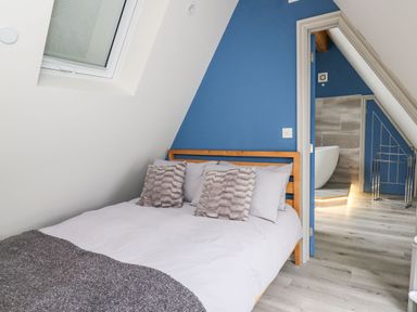 A bedroom with a wooden bed with pillows and a blanket next to a window and a door leading to a bathroom at Hill Radnor - The Sheepfold in Llanyre near Llandrindod Wells