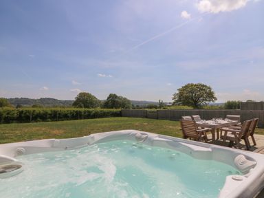 An outdoor hot tub with bubbling water and wooden dining table with chairs on a patio in a grassy yard at Hill Radnor - The Sheepfold in Llanyre near Llandrindod Wells