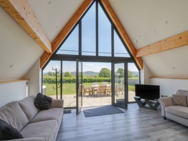 A living room with sofas a TV on a stand a coat rack and large glass doors opening to a patio with outdoor furniture at Hill Radnor - The Sheepfold in Llanyre near Llandrindod Wells