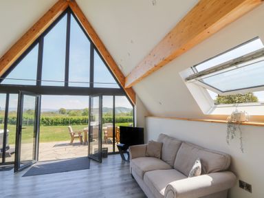 A living room with a beige sofa a television and large triangular windows overlooking an outdoor patio with chairs at Hill Radnor - The Sheepfold in Llanyre near Llandrindod Wells