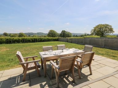 A wooden outdoor dining table with six chairs on a stone patio in a garden with trees and hedges at Hill Radnor - The Sheepfold in Llanyre near Llandrindod Wells