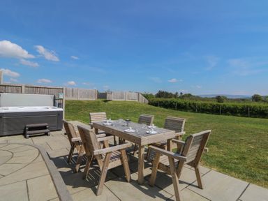 An outdoor patio with a wooden dining table and six chairs set with plates and cups near a hot tub at Hill Radnor - The Sheepfold in Llanyre near Llandrindod Wells