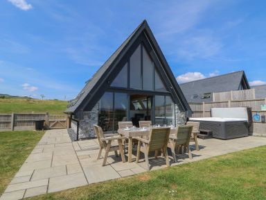 A patio with outdoor wooden dining furniture and a hot tub next to a modern A-frame house at Hill Radnor - The Sheepfold in Llanyre near Llandrindod Wells