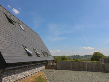 Exterior view of a building with gray roof shingles and several open windows next to a gravel area and wooden fence with trees and hills in the background at Hill Radnor - The Sheepfold in Llanyre near Llandrindod Wells