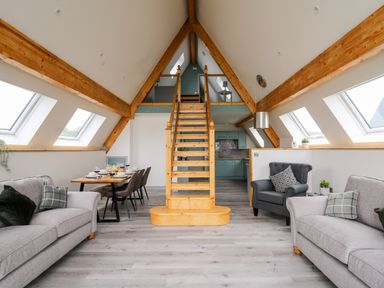 A living room with gray sofas a gray armchair a dining table with chairs and a wooden staircase leading to a loft at Brecknock Cheviot - The Sheepfold in Llanyre near Llandrindod Wells