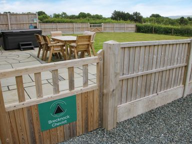 An outdoor patio area with wooden table and chairs next to a hot tub enclosed by wooden fencing at Brecknock Cheviot - The Sheepfold in Llanyre near Llandrindod Wells