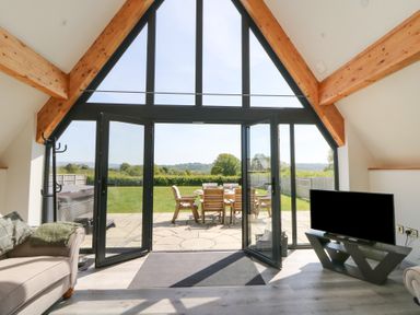 A room with a sofa a television and large windows overlooking a patio and garden at Brecknock Cheviot - The Sheepfold in Llanyre near Llandrindod Wells