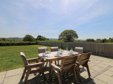 A wooden outdoor dining table with six chairs set with dishes and glasses on a stone patio overlooking a fenced grassy yard at Brecknock Cheviot - The Sheepfold in Llanyre near Llandrindod Wells