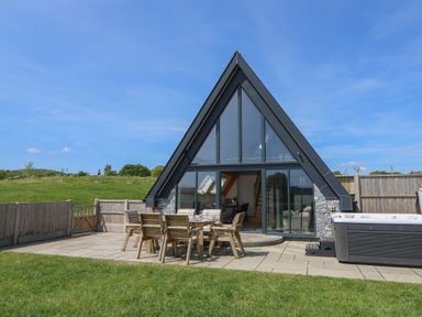 An outdoor patio with wooden table and chairs next to a modern a frame house with large windows and a hot tub at Brecknock Cheviot - The Sheepfold in Llanyre near Llandrindod Wells