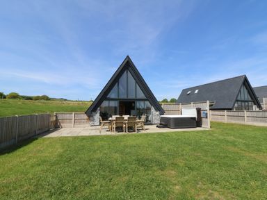 A fenced outdoor area with wooden chairs and a table and a hot tub in front of a triangular house at Brecknock Cheviot - The Sheepfold in Llanyre near Llandrindod Wells