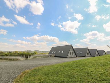 A row of triangular stone and tile roof buildings on a gravel area with a wooden fence and grassy hill at Brecknock Cheviot - The Sheepfold in Llanyre near Llandrindod Wells