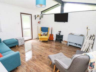 A living room with a blue sofa a yellow armchair a grey armchair with footstool a wall mounted television and a guitar at Doirewood in Penlan Holiday Park near Cenarth