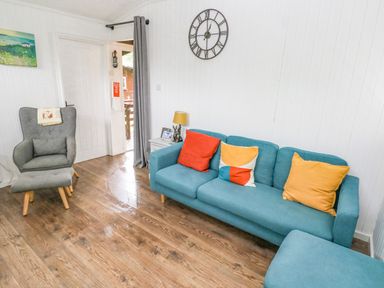 A living room with a blue sofa with cushions a grey armchair with footstool a side table with a lamp and wall clock at Doirewood Penlan Holiday Park near Cenarth