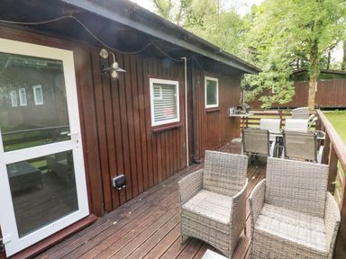 An outdoor wooden deck attached to a cabin with wicker chairs a dining table and barbecue grill at Doirewood Penlan Holiday Park near Cenarth