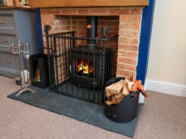 A wood-burning stove with a fire inside surrounded by a black metal safety gate with firewood and fireplace tools nearby at Over Esk in Whitby