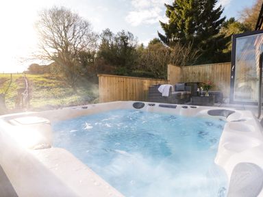 An outdoor hot tub with bubbling water next to a patio with wicker seating and a table with flowers at The Linhay Cadeleigh near Tiverton