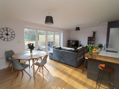A living room with a gray sofa a round wooden dining table with chairs and a kitchen counter with bar stools at The Linhay in Cadeleigh near Tiverton