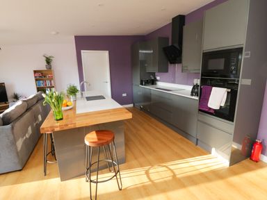 A kitchen with grey cabinets wooden stools a white countertop and a grey sofa at The Linhay in Cadeleigh near Tiverton