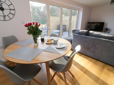A dining area with a wooden round table with grey chairs and a sofa with a television in a living room at The Linhay in Cadeleigh near Tiverton