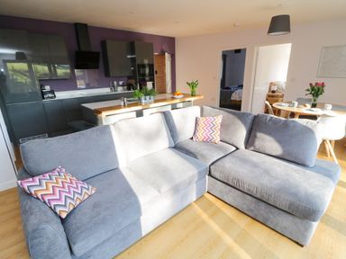 A living room with a gray sectional sofa and colorful pillows next to a kitchen with dark cabinets and a dining area with a round table and chairs at The Linhay in Cadeleigh near Tiverton