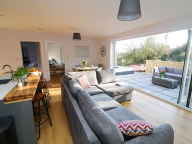 A living room with a gray sectional sofa and a kitchen island with bar stools next to a dining area and a patio with outdoor seating at The Linhay in Cadeleigh near Tiverton