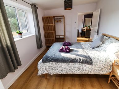 A bedroom with a double bed, a wooden open wardrobe, a window with curtains, and a bedside table at The Linhay in Cadeleigh near Tiverton