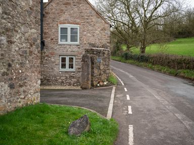 A stone cottage with two windows next to a curved road and grassy area with a large rock at Wavering in Compton Bishop near Axbridge