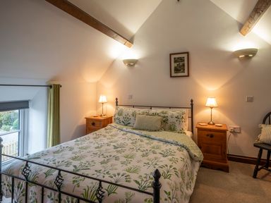A bedroom with a bed with floral bedding two bedside tables with lamps a window with curtains and wooden beams on the ceiling at Crooks in Axbridge