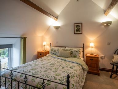 A bedroom with a double bed with floral bedding two wooden bedside tables with lamps a window with a green curtain and a framed picture on the wall at Crooks in Axbridge