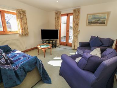 A sitting room with blue upholstered chairs a television on a stand a small wooden table with a board game and floral curtains at Granary Cottage in Blackwell near Ilmington